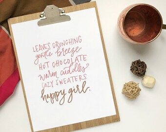 May include: A wooden clipboard with a white paper featuring handwritten text in pink and gold: "Leaves crunching, gentle breeze, hot chocolate, warm cuddles, cozy sweaters, happy girl." A copper mug and decorative spheres are also present.