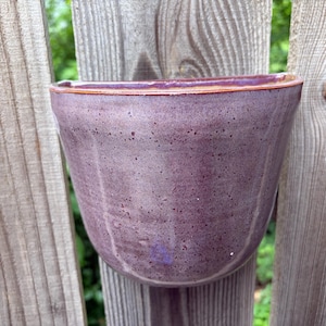May include: A wall-mounted ceramic planter with a semi-circular shape and a flat back. The exterior features a speckled purple glaze with subtle vertical indentations along the body and a smooth, rounded rim with a brownish-orange edge finish.