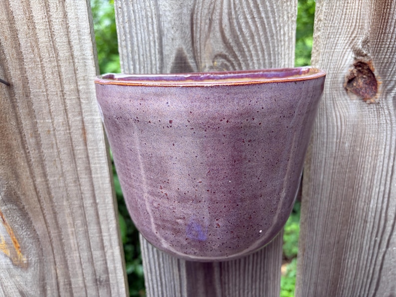 May include: A wall-mounted ceramic planter with a semi-circular shape and a flat back. The exterior features a speckled purple glaze with subtle vertical indentations along the body and a smooth, rounded rim with a brownish-orange edge finish.