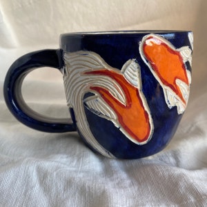 Pu&ograve; includere: Una tazza in ceramica con una glassa blu scuro. La tazza presenta un disegno di carpe koi arancioni e bianche. Il manico &egrave; anch'esso blu scuro. Le carpe koi sono su uno sfondo di motivi bianchi a forma di onda.