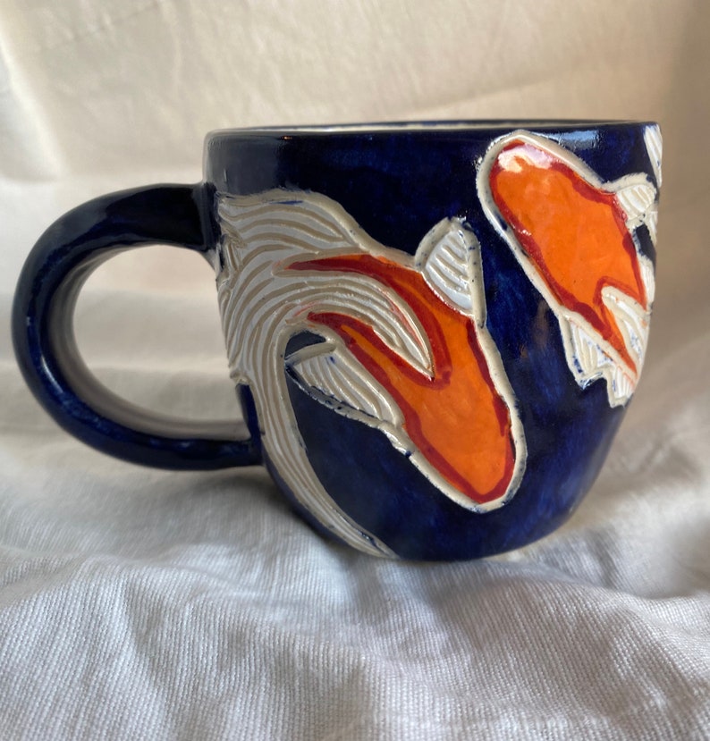 Pu&ograve; includere: Una tazza in ceramica con una glassa blu scuro. La tazza presenta un disegno di carpe koi arancioni e bianche. Il manico &egrave; anch'esso blu scuro. Le carpe koi sono su uno sfondo di motivi bianchi a forma di onda.