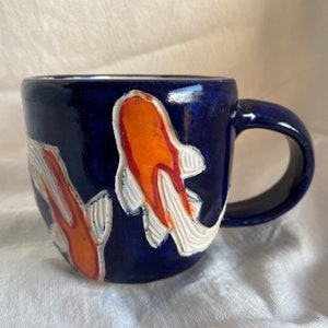 Pu&ograve; includere: Una tazza in ceramica con smalto blu scuro. La tazza presenta due disegni di pesci koi arancioni e bianchi. Il manico &egrave; anch'esso blu scuro. La tazza &egrave; progettata per bevande calde o fredde.