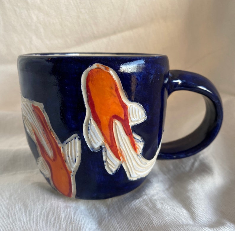 Pu&ograve; includere: Una tazza in ceramica con smalto blu scuro. La tazza presenta due disegni di pesci koi arancioni e bianchi. Il manico &egrave; anch'esso blu scuro. La tazza &egrave; progettata per bevande calde o fredde.