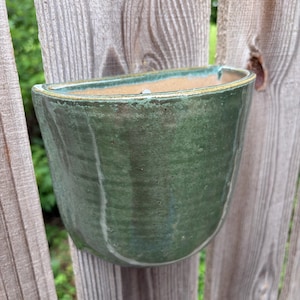 May include: A semi-circular wall-mounted ceramic planter featuring a glossy, mottled forest green glaze with subtle vertical texture. The interior of the vessel reveals a natural, unglazed tan clay finish, and the top rim is accented with a thin, golden-brown edge.
