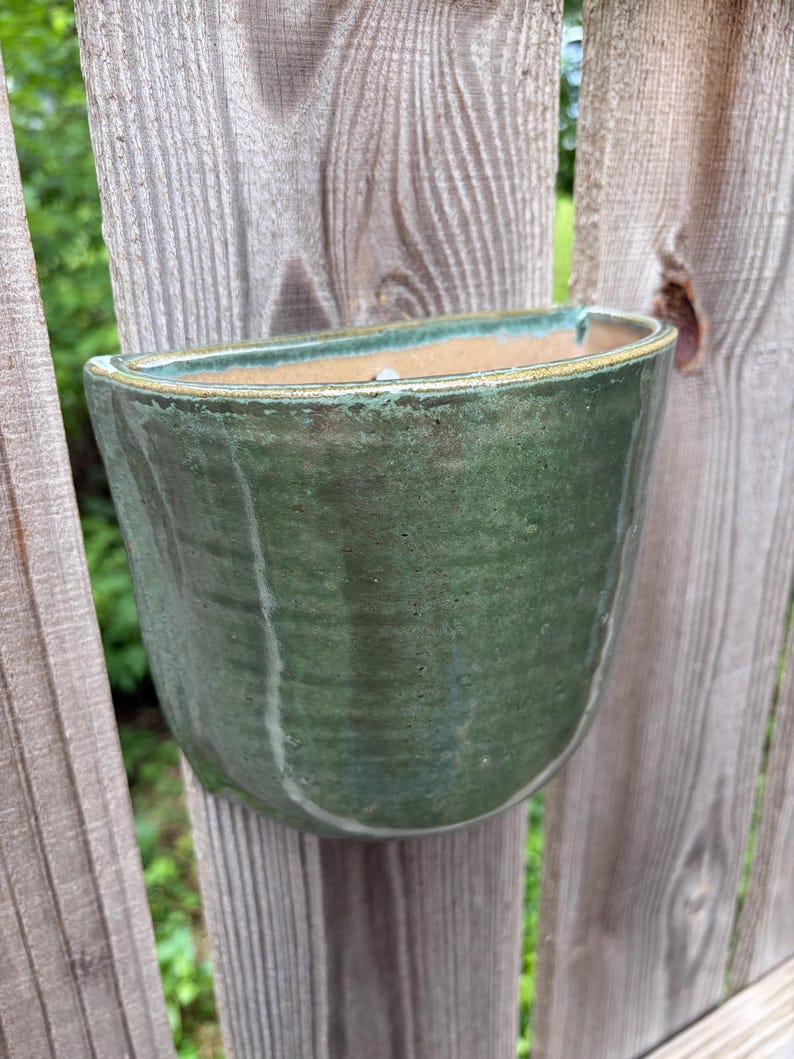 May include: A semi-circular wall-mounted ceramic planter featuring a glossy, mottled forest green glaze with subtle vertical texture. The interior of the vessel reveals a natural, unglazed tan clay finish, and the top rim is accented with a thin, golden-brown edge.