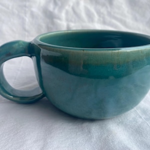 Peut inclure: Une tasse en céramique turquoise avec un corps arrondi et une anse incurvée. La tasse a une finition brillante avec des variations de couleur, passant d'un turquoise plus foncé sur le bord à un bleu-turquoise plus clair sur le corps. La tasse est conçue pour les boissons.