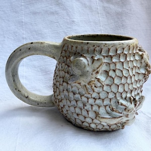 May include: A ceramic mug with a speckled off-white glaze featuring a raised honeycomb texture across the exterior. The mug includes a large curved handle and sculpted bee figures integrated into the hexagonal pattern on the side of the vessel.