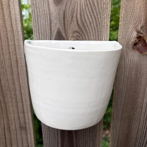 May include: A semi-circular white ceramic wall planter with a smooth, glossy finish and a flat back. The vessel features a slightly irregular, hand-crafted texture along its curved front surface and a small hole at the top for mounting purposes.