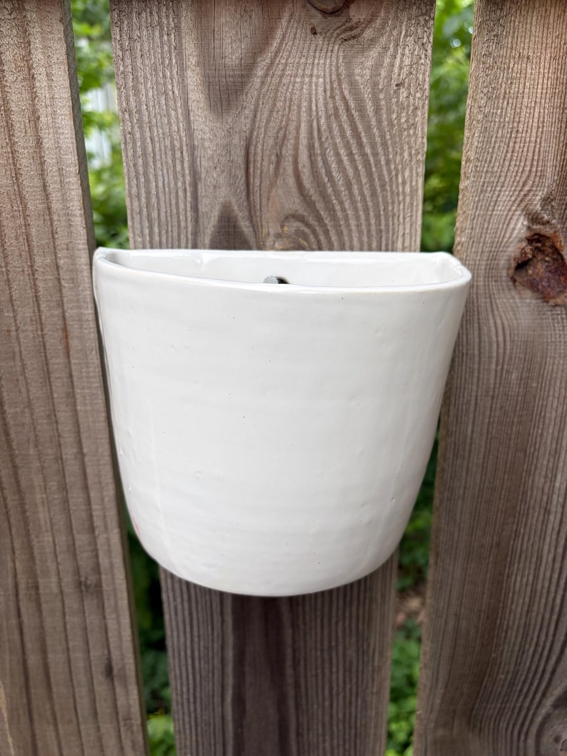 May include: A semi-circular white ceramic wall planter with a smooth, glossy finish and a flat back. The vessel features a slightly irregular, hand-crafted texture along its curved front surface and a small hole at the top for mounting purposes.