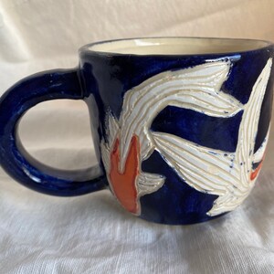 Pu&ograve; includere: Una tazza in ceramica con esterno blu scuro e un design a carpa koi bianco e arancione. La tazza ha un manico arrotondato e un interno color crema. Il design della carpa koi &egrave; in rilievo e strutturato.