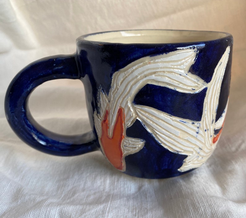 Pu&ograve; includere: Una tazza in ceramica con esterno blu scuro e un design a carpa koi bianco e arancione. La tazza ha un manico arrotondato e un interno color crema. Il design della carpa koi &egrave; in rilievo e strutturato.
