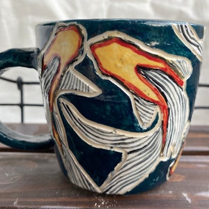 Pu&ograve; includere: Una tazza in ceramica con smalto blu scuro e un design strutturato. Il design presenta forme astratte gialle e rosse con accenti a righe bianche e nere. La tazza ha un manico arrotondato ed &egrave; adatta per bevande calde.