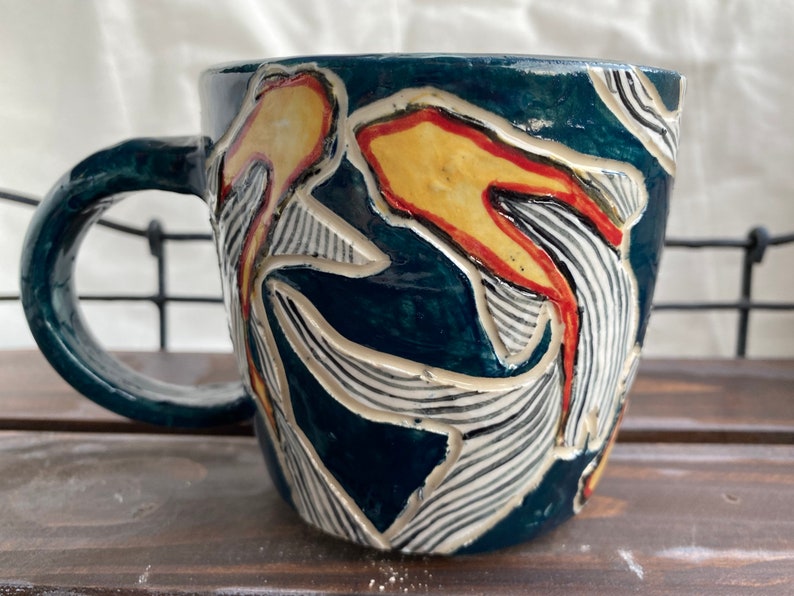 Pu&ograve; includere: Una tazza in ceramica con smalto blu scuro e un design strutturato. Il design presenta forme astratte gialle e rosse con accenti a righe bianche e nere. La tazza ha un manico arrotondato ed &egrave; adatta per bevande calde.