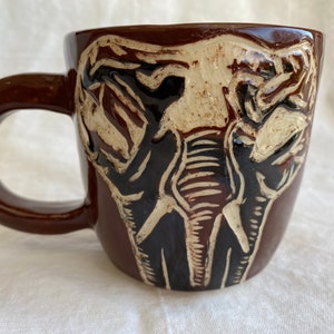 May include: A brown ceramic mug with a raised design of an elephant's face. The elephant's face is in shades of brown, black, and white.