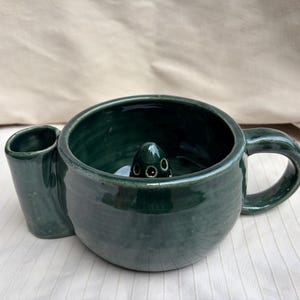 May include: A dark green ceramic shaving bowl with a glossy finish, featuring a side handle, a cylindrical brush holder attachment, and a central raised dome with small holes designed for lathering soap during the shaving process.