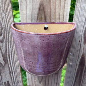 May include: A semi-circular ceramic wall planter with a glossy purple exterior glaze featuring subtle dark speckles. The interior of the vessel is a smooth, light tan color, and a single circular hole is centered near the top edge for mounting.