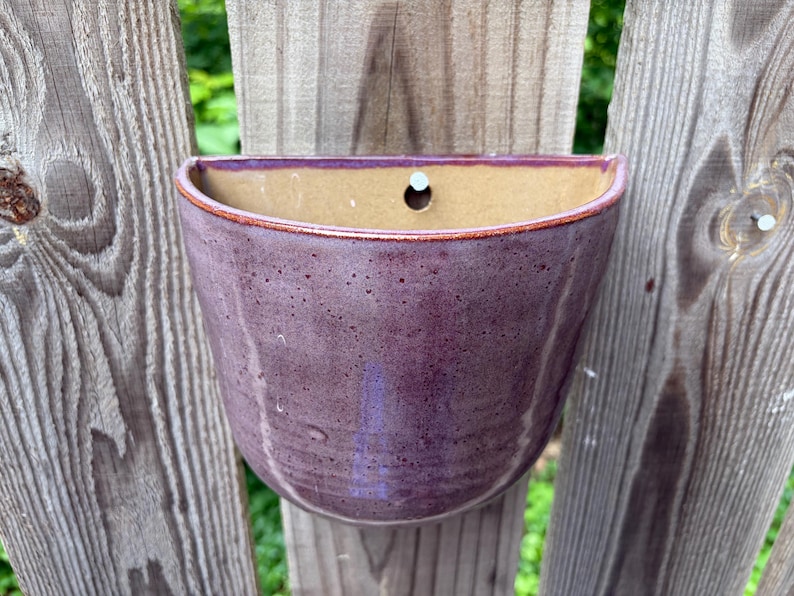 May include: A semi-circular ceramic wall planter with a glossy purple exterior glaze featuring subtle dark speckles. The interior of the vessel is a smooth, light tan color, and a single circular hole is centered near the top edge for mounting.