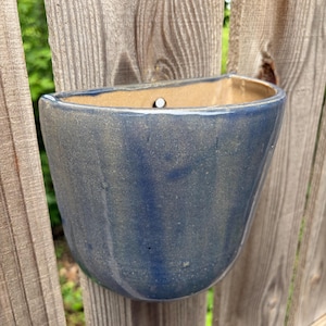 May include: A semi-circular wall-mounted ceramic planter with a glossy, deep blue glaze and a smooth, curved exterior. The interior of the planter is a light tan color and features a single circular hole at the top center for mounting purposes.