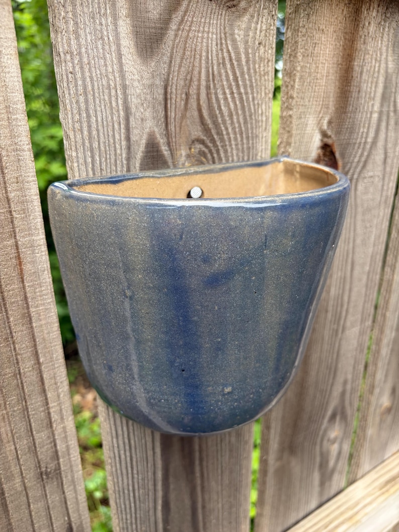 May include: A semi-circular wall-mounted ceramic planter with a glossy, deep blue glaze and a smooth, curved exterior. The interior of the planter is a light tan color and features a single circular hole at the top center for mounting purposes.