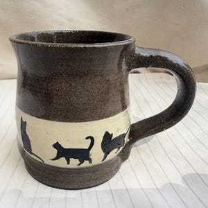 May include: A ceramic mug with a dark brown speckled glaze on the top and bottom sections. The center features a wide, cream-colored band decorated with three black cat silhouettes in various poses, including one sitting and two walking.