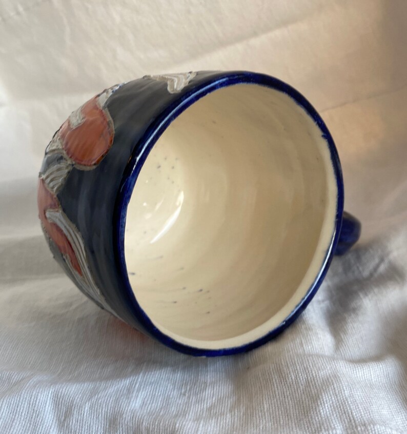 Pu&ograve; includere: Una tazza in ceramica con esterno blu scuro e interno color crema. La tazza ha un manico ed &egrave; decorata con disegni floreali rossi e bianchi. Il bordo e il manico sono anch'essi blu scuro.