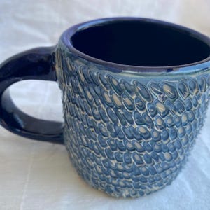 May include: A handmade ceramic mug with a textured, scale-like pattern in shades of blue and white. The mug has a dark blue interior and handle. This unique mug is perfect for coffee or tea.
