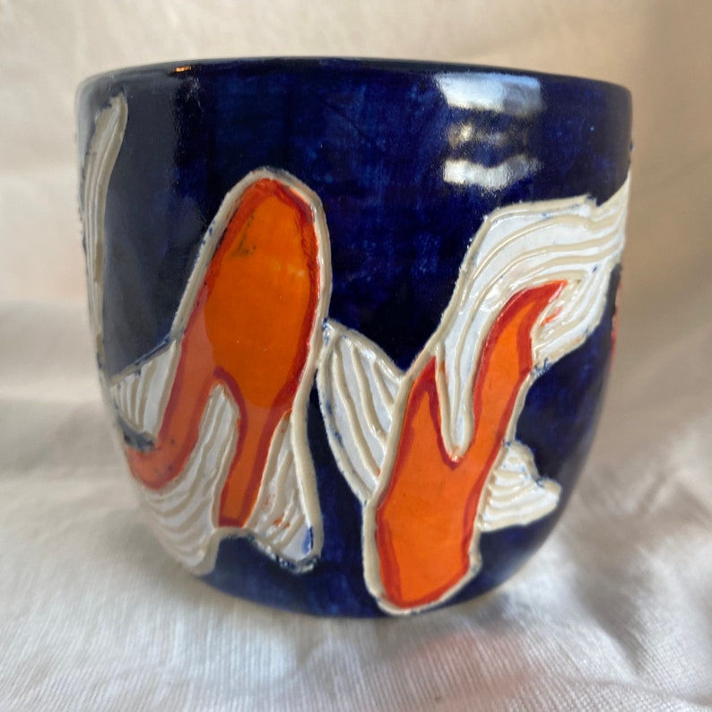 Pu&ograve; includere: Una tazza in ceramica con una glassa blu scuro, con disegni di pesci koi arancioni con accenti bianchi. La tazza ha una forma arrotondata ed &egrave; probabilmente fatta a mano, mostrando uno stile artistico unico.