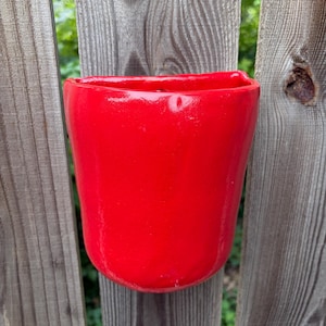 May include: A bright red, semi-circular wall-mounted planter with a smooth, glossy finish. The vessel features a flat back edge designed for flush mounting against a vertical surface, with a wide, open top and a rounded, slightly irregular base.
