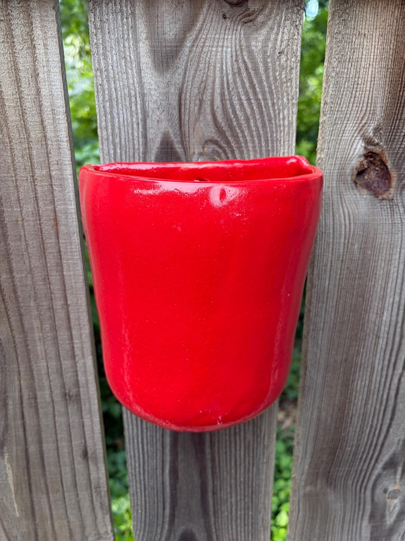 May include: A bright red, semi-circular wall-mounted planter with a smooth, glossy finish. The vessel features a flat back edge designed for flush mounting against a vertical surface, with a wide, open top and a rounded, slightly irregular base.