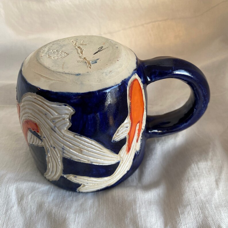 Pu&ograve; includere: Una tazza in ceramica con smalto blu scuro e interno bianco. La tazza presenta un disegno intagliato di pesci koi con dettagli arancioni e bianchi. Il manico &egrave; blu scuro. Il fondo della tazza ha un marchio del produttore.