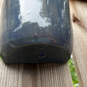 May include: A semi-circular ceramic wall planter with a glossy, deep navy blue glaze. The surface features subtle speckling and a smooth, reflective finish. A single circular mounting hole is centered near the bottom edge of the curved base.