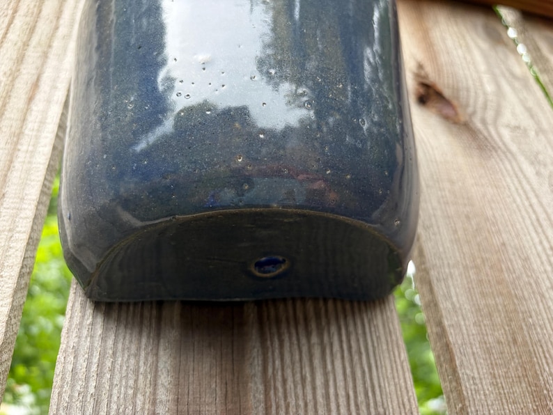 May include: A semi-circular ceramic wall planter with a glossy, deep navy blue glaze. The surface features subtle speckling and a smooth, reflective finish. A single circular mounting hole is centered near the bottom edge of the curved base.