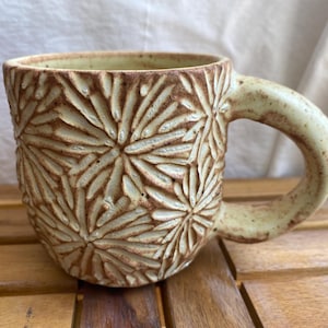 May include: A ceramic mug with a textured floral design. The mug is light beige with brown detailing and a rounded handle. It sits on a wooden surface. The mug is suitable for hot drinks.