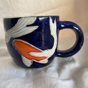 Puede incluir: Una taza de cerámica con un esmalte azul oscuro. La taza presenta un diseño pintado a mano de peces koi en naranja y blanco. El asa es un lazo redondeado y la taza se encuentra sobre una superficie blanca.