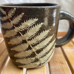 Puede incluir: Una taza de cerámica hecha a mano con un esmalte marrón oscuro y un diseño de hoja de helecho color crema. La taza tiene un asa redondeada y una forma cuadrada. Esta taza única es perfecta para café, té o cualquier bebida.