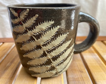 May include: A handmade ceramic mug with a dark brown glaze and a cream-colored fern leaf design. The mug has a rounded handle and is approximately 4 inches tall.
