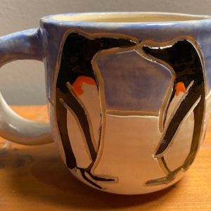May include: A ceramic mug with a blue and white glaze. The mug features two penguins in black and white with orange accents. The penguins are facing each other and appear to be interacting.