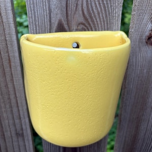 May include: A bright yellow ceramic wall planter with a textured, slightly pitted surface. The vessel features a semi-circular opening at the top and a single circular hole near the upper rim, designed for mounting against a vertical surface.