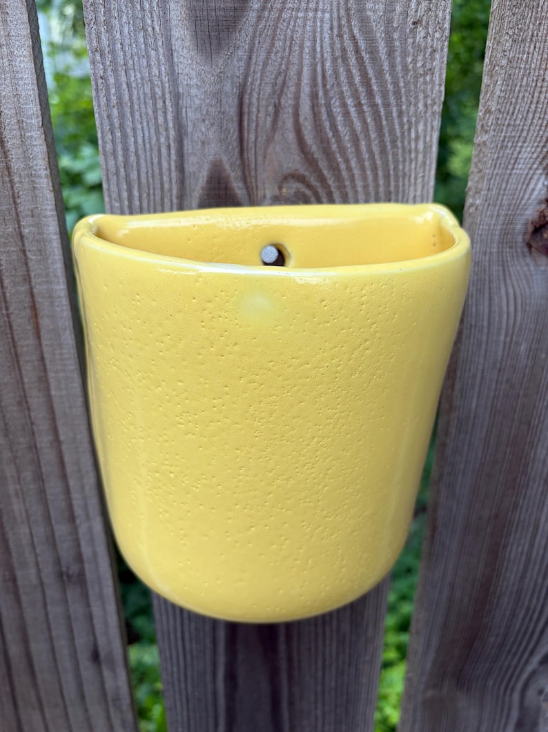 May include: A bright yellow ceramic wall planter with a textured, slightly pitted surface. The vessel features a semi-circular opening at the top and a single circular hole near the upper rim, designed for mounting against a vertical surface.