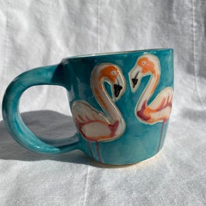 Op de afbeelding: Een turquoise keramische mok met een gebogen handvat. De mok heeft twee flamingo-illustraties in oranje, roze en wit. Een uniek stuk drinkgerei, perfect voor koffie, thee of andere dranken.