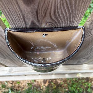 May include: A semi-circular ceramic wall planter with a glossy brown interior and a dark, mottled exterior finish. The vessel features a flat back edge with a pre-drilled hole for mounting and a drainage hole at the bottom of the basin.