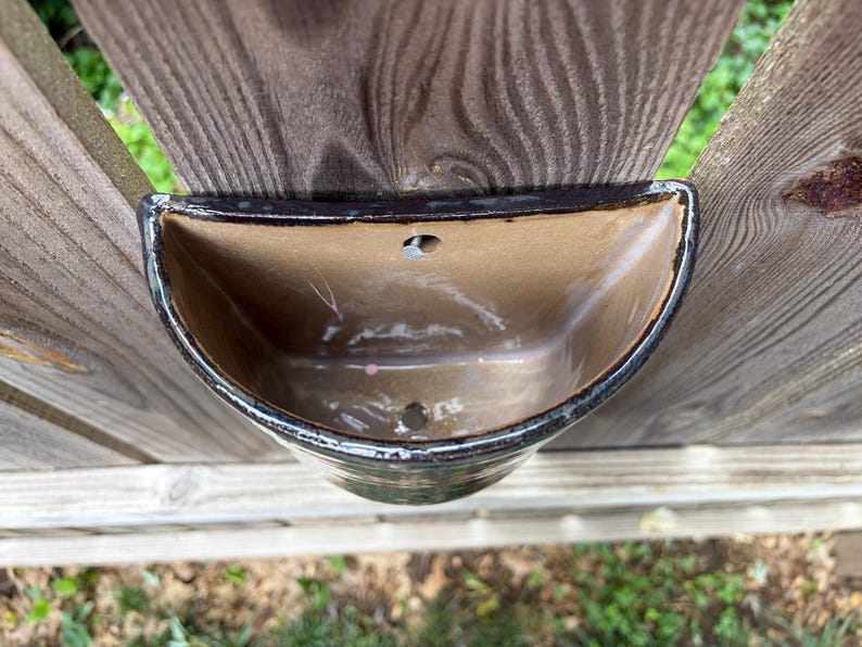 May include: A semi-circular ceramic wall planter with a glossy brown interior and a dark, mottled exterior finish. The vessel features a flat back edge with a pre-drilled hole for mounting and a drainage hole at the bottom of the basin.