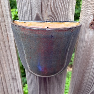 May include: A semi-circular wall-mounted ceramic planter featuring a glossy glaze with blended tones of deep blue, charcoal, and hints of reddish-brown. The interior of the vessel has a smooth, light tan finish with a visible drainage hole.