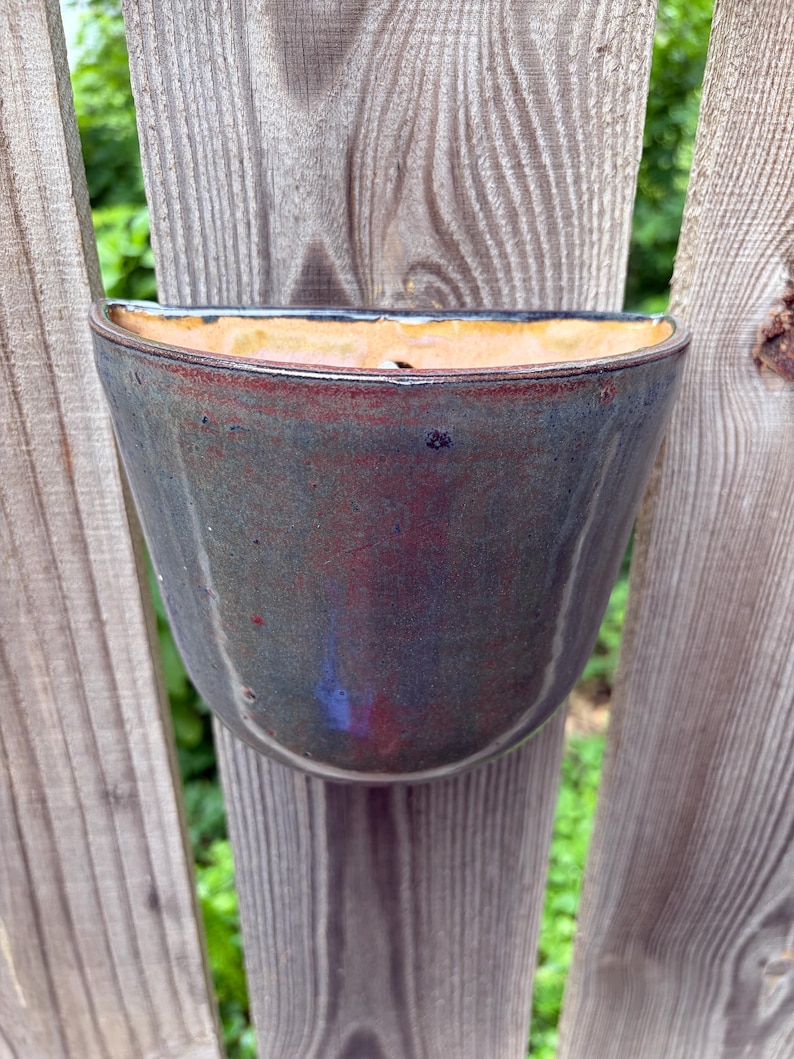 May include: A semi-circular wall-mounted ceramic planter featuring a glossy glaze with blended tones of deep blue, charcoal, and hints of reddish-brown. The interior of the vessel has a smooth, light tan finish with a visible drainage hole.