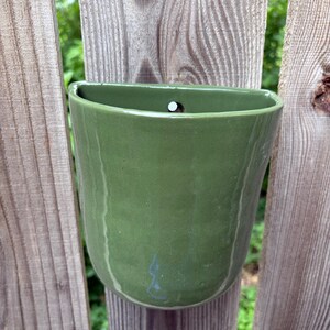 May include: A wall-mounted ceramic planter with a smooth, sage green glaze. The vessel features a semi-circular opening at the top with a small, centered hole for hanging. The surface has a subtle, organic texture with slight color variations.