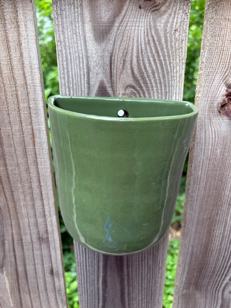 May include: A wall-mounted ceramic planter with a smooth, sage green glaze. The vessel features a semi-circular opening at the top with a small, centered hole for hanging. The surface has a subtle, organic texture with slight color variations.
