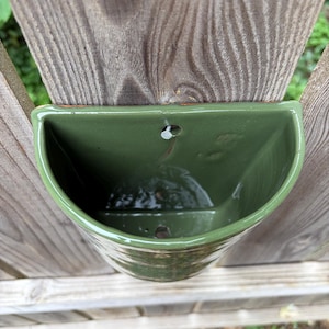 May include: A semi-circular wall-mounted planter with a glossy olive green glaze. The ceramic vessel features a flat back edge with a pre-drilled hole for mounting and a drainage hole at the bottom, showcasing a smooth, reflective surface finish.