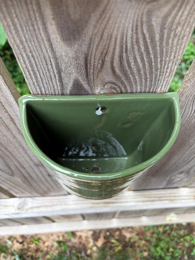 May include: A semi-circular wall-mounted planter with a glossy olive green glaze. The ceramic vessel features a flat back edge with a pre-drilled hole for mounting and a drainage hole at the bottom, showcasing a smooth, reflective surface finish.