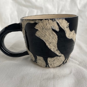 May include: Black ceramic mug with a carved crow design. The mug has a black handle and a cream interior. The exterior features a textured, carved relief of crows against a black background. A unique, handmade piece.