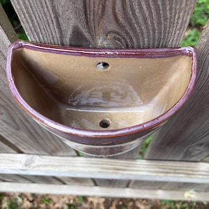 May include: A semi-circular wall-mounted ceramic planter with a glossy tan interior and a dark reddish-brown rim. The piece features a flat back with a pre-drilled hole for mounting and a drainage hole at the bottom of the basin for plant care.